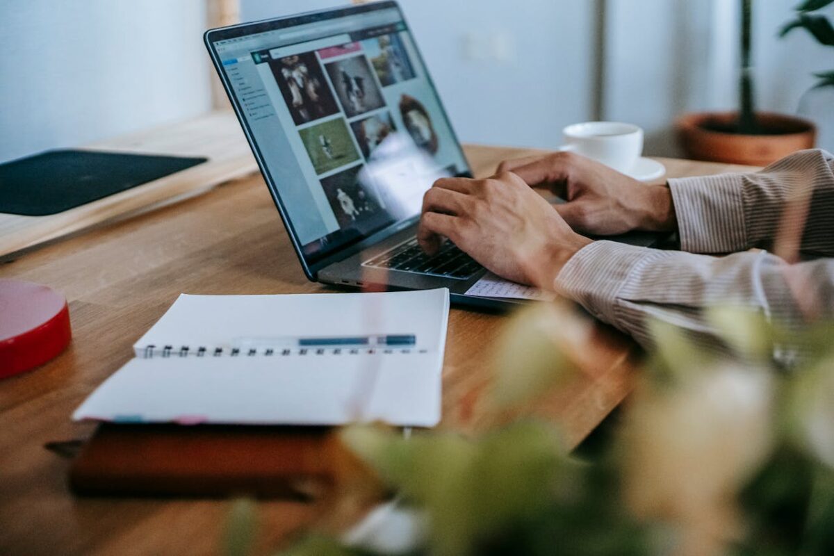 Crop unrecognizable guy in casual outfit working remotely on computer while sitting at wooden table near notebook with cup of coffee in light room